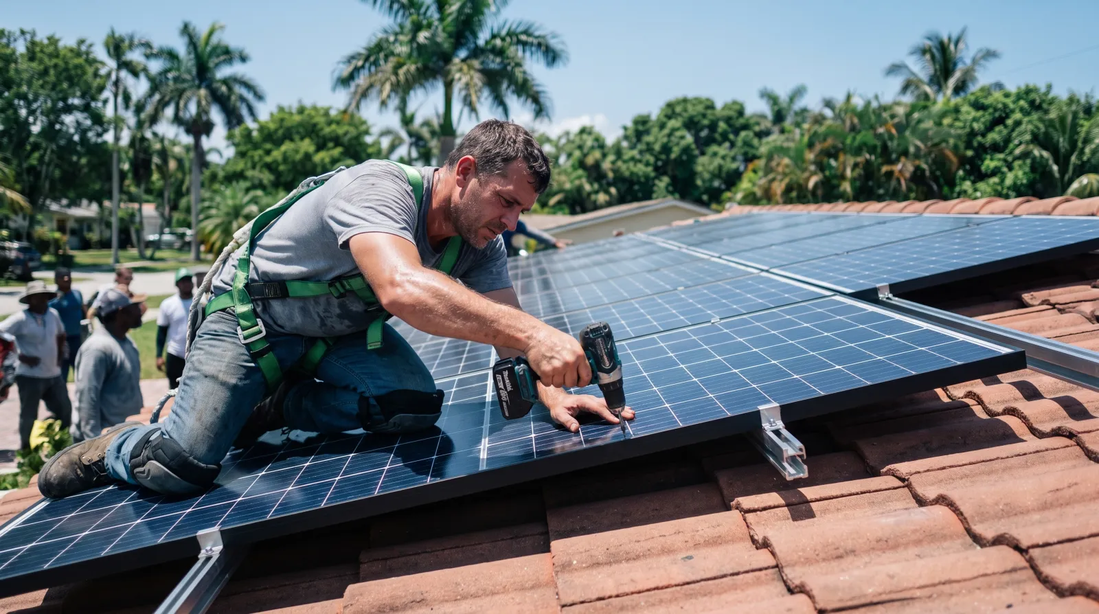 A solar installer kneels on a tile roof in South Florida, securing a photovoltaic panel into place with a drill.