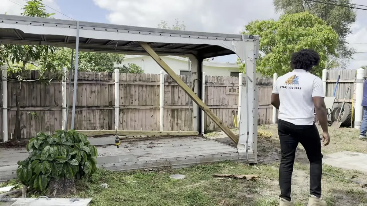 Rapid Haul crew member in a company shirt walking up to a backyard shed and carport before the tear-down