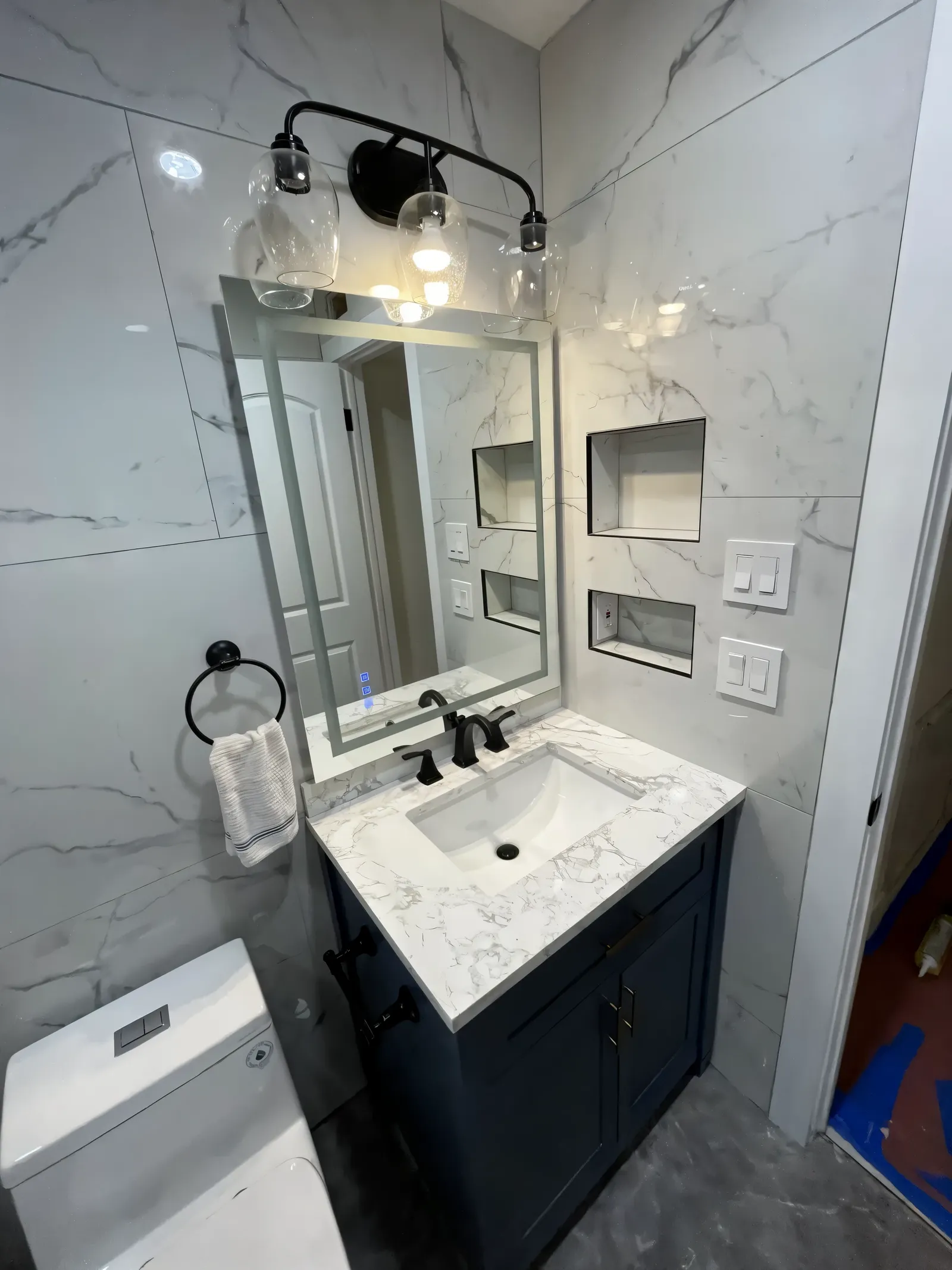 The same bathroom after the remodel with marble-look tile, a navy vanity, black fixtures and a lit mirror